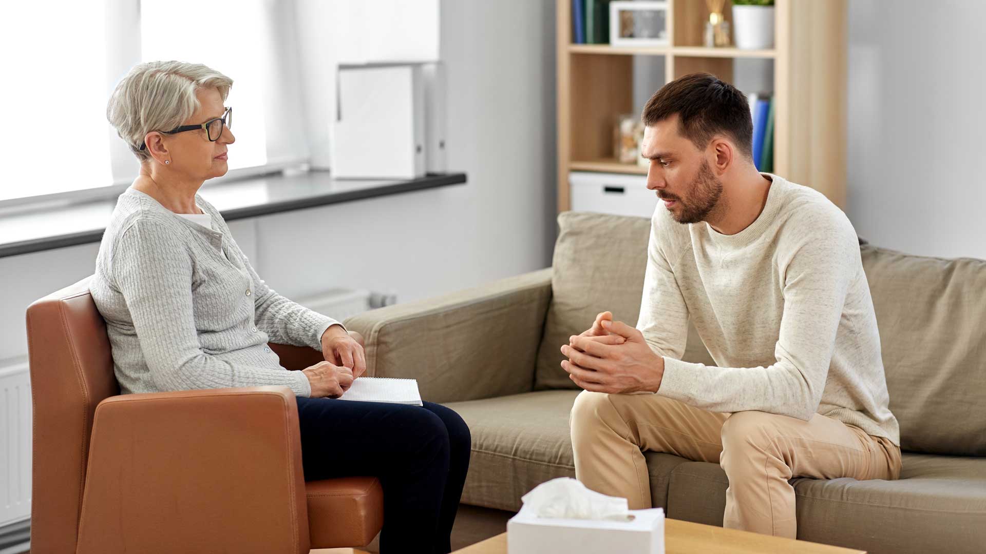 Eine Frau und ein Mann sitzen in einem Gesprächssetting mit Sofa, Sessel und Tisch in einem hellen Raum.
