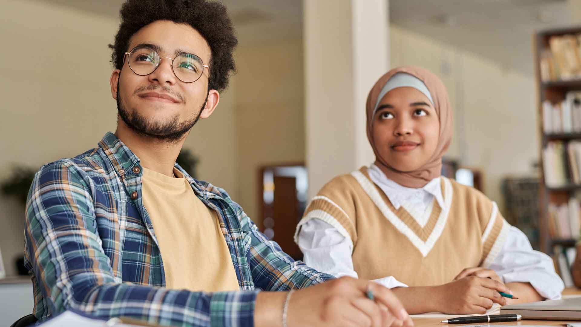 Ein junger Mann mit Brille blickt zur Seite, eine Frau mit Kopftuch sitzt daneben.